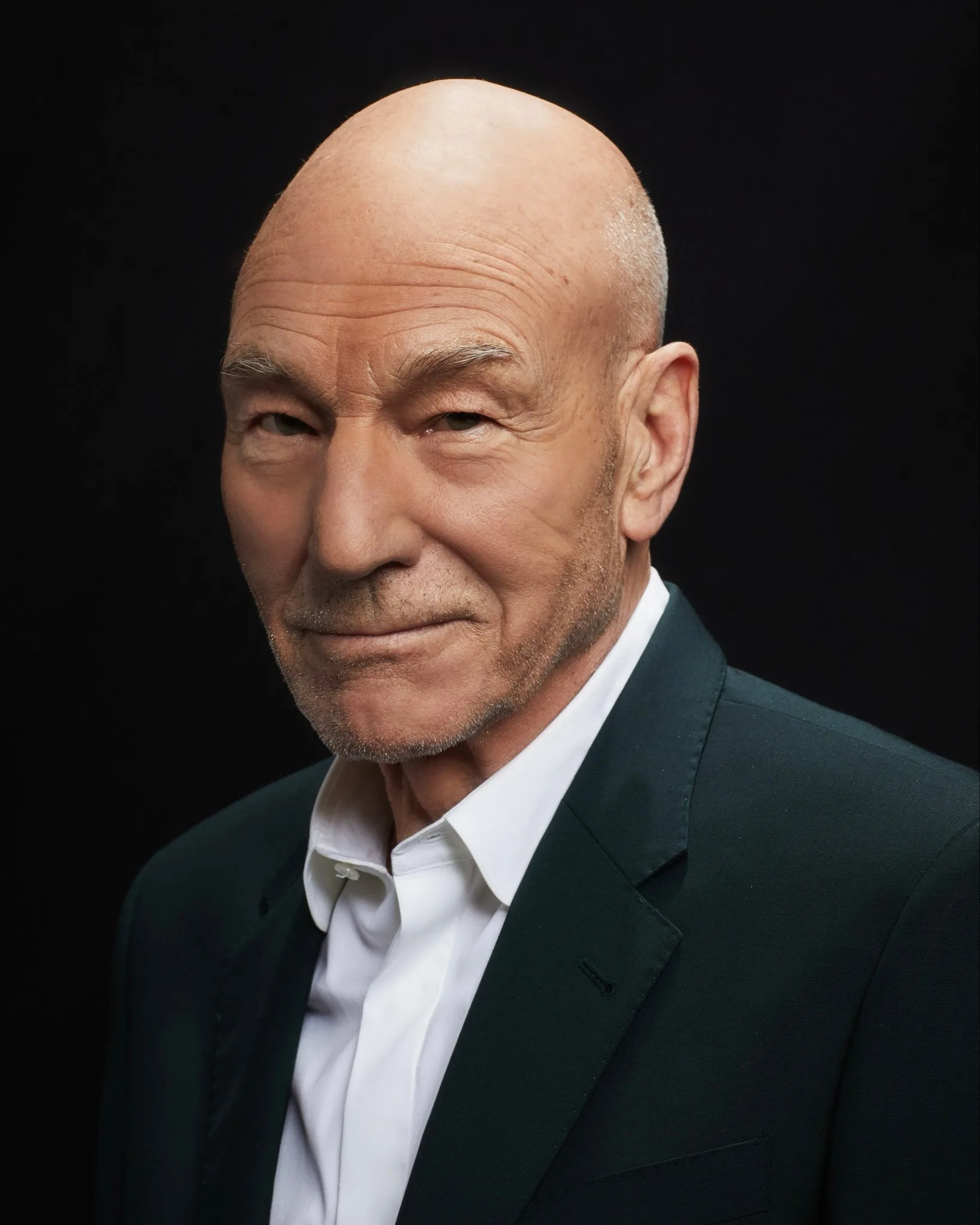 Portrait of Sir Patrick Stewart in a dark jacket and white shirt against a black background.
