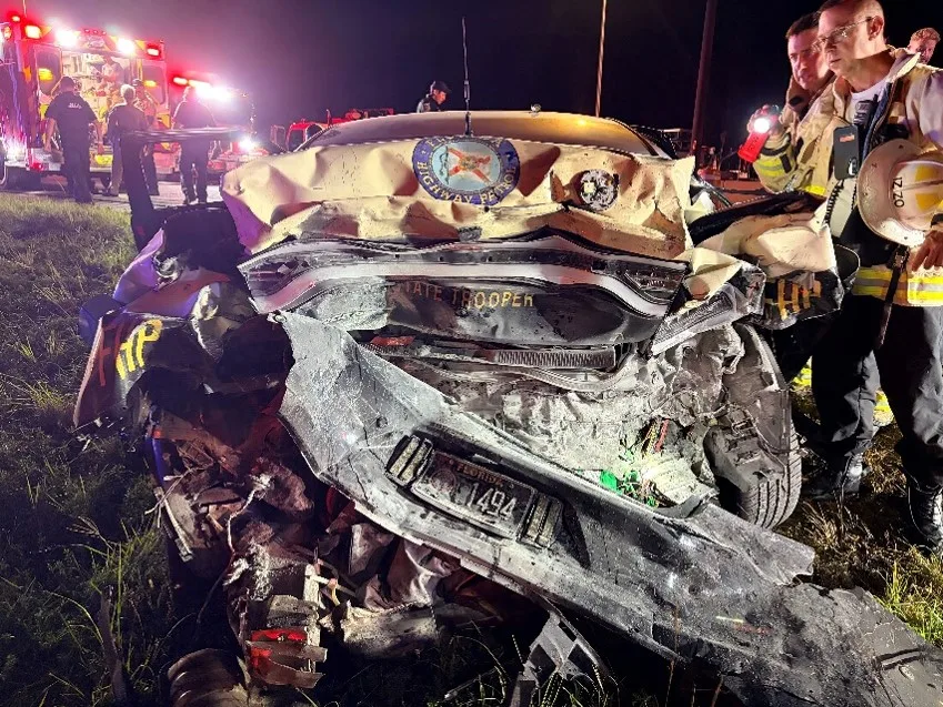 The heavily damaged rear of a Florida Highway Patrol car at night after a crash on State Road 429, with emergency vehicle lights in the background.
